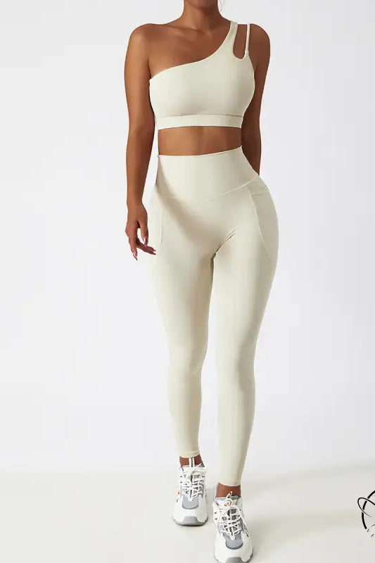 Cream-colored one-shoulder sports bra and high-waisted yoga pants set with white sneakers