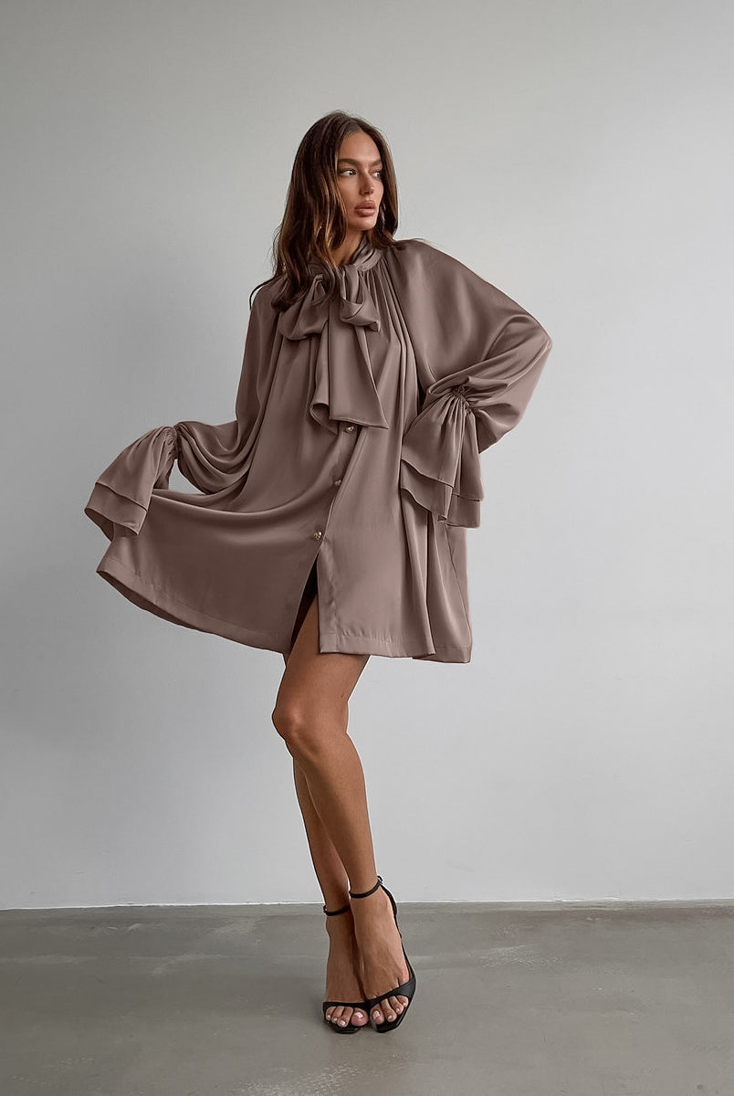 Flowy taupe satin dress features oversized bow and billowing sleeves for elegant fashion
