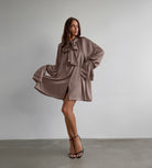 Flowy taupe satin dress features oversized bow and billowing sleeves for elegant fashion