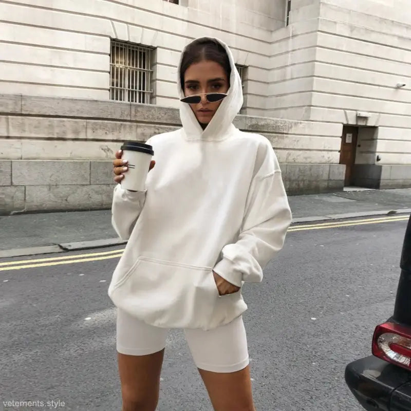 Woman in white hoodie and shorts holding coffee and sunglasses, showcasing army green sports sweater suit