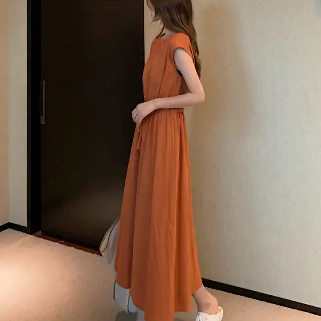 Long orange sleeveless dress with a flowy skirt worn over light-colored pants and white sneakers.