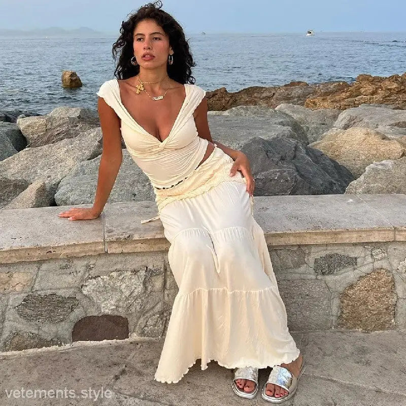 Woman in a flowing white dress sitting on a rocky coastal ledge showcasing the elegant low cut short French outdoor set