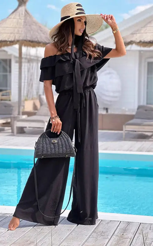 Woman in black off shoulder jumpsuit and straw hat, featuring elegant ruffle design