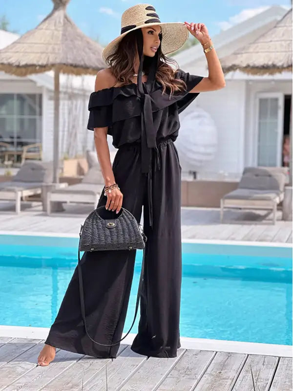 Woman in black off shoulder jumpsuit and straw hat, featuring elegant ruffle design