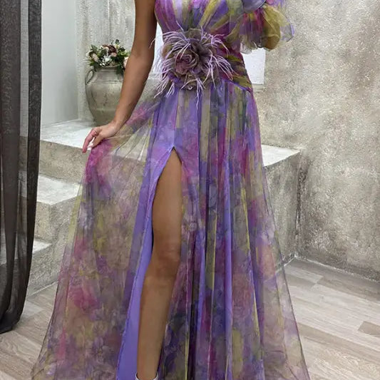 Flowing one-shoulder gown in sheer purple, pink, and gold tones with a high slit and feathered waist detail.