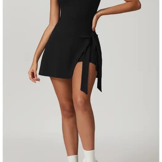 Black sleeveless mini dress with a side slit and tie detail, paired with white socks and chunky sneakers.
