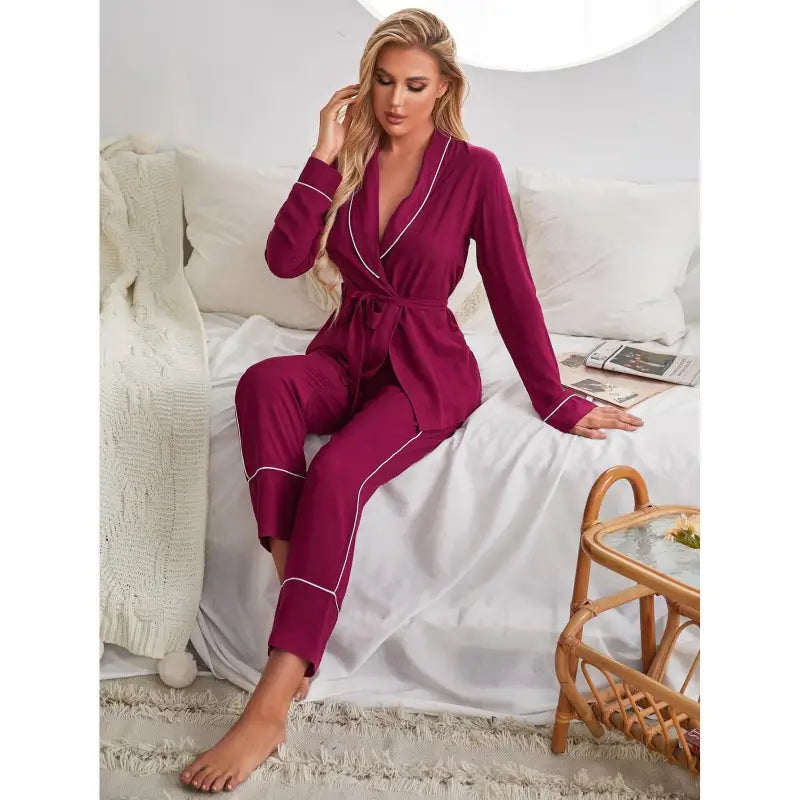Blonde woman seated on bed wearing magenta satin pajamas with white piping, ideal as winter red cardigan sleepwear