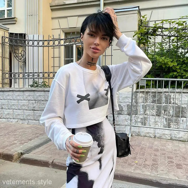 Woman in white long sleeve pullover with black X graphic and matching skirt holding coffee