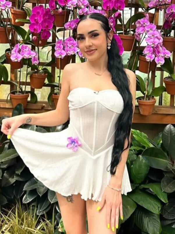 Woman in a white strapless mini dress posing among orchid plants.